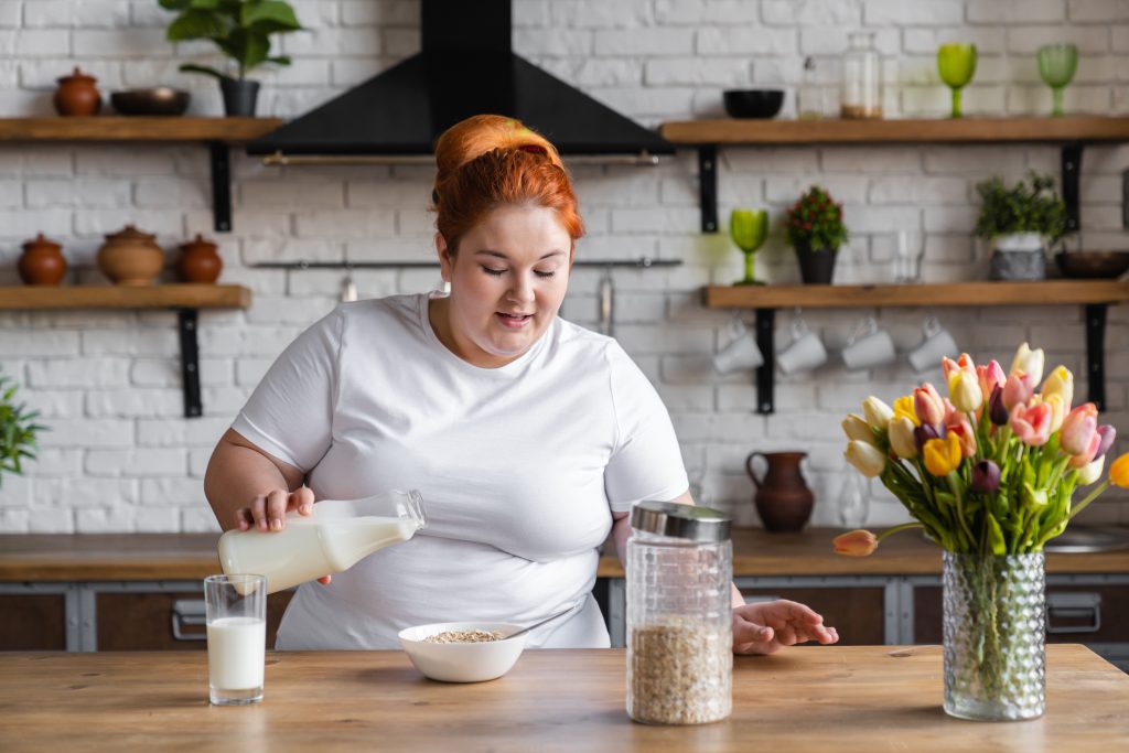 Young chubby woman standing in kitchen pouring milk into oat-flakes smiling